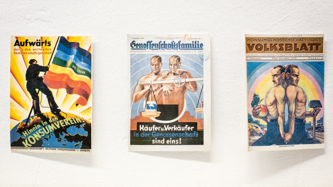 Drei Zeitschriftentitel an die Wand gepinnt. Auf dem mittleren zwei wie Zwillinge aussehende Männer hinter einer Pendelwaage, darunter geschrieben steht: "Käufer und Verkäufer in der Genossenschaft sind eins!"