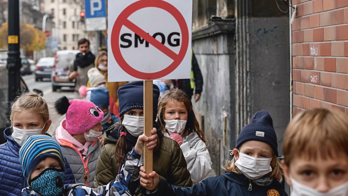 Kinder mit Atemschutzmasken stehen auf einer Straße in Katowice, zwei von ihnen halten Protestschild Smog hoch