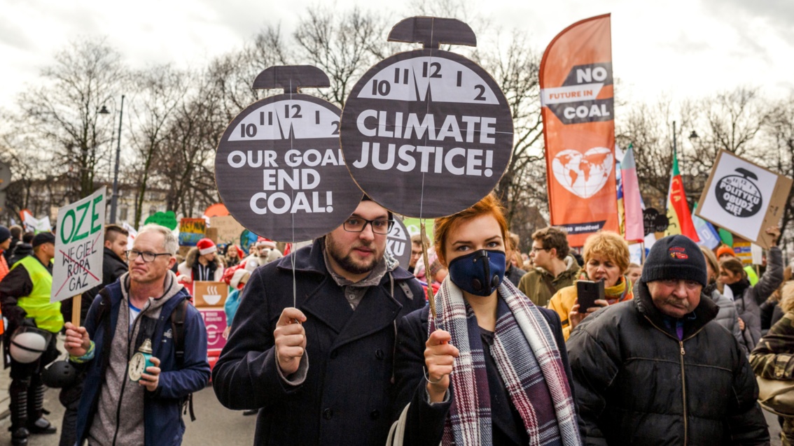 Ein Paar hält jeweils ein Schild in Form eines Weckers, oben die Uhr, unten die Aufschrift: "Climate Justice" und "Our goal is End of Coal"
