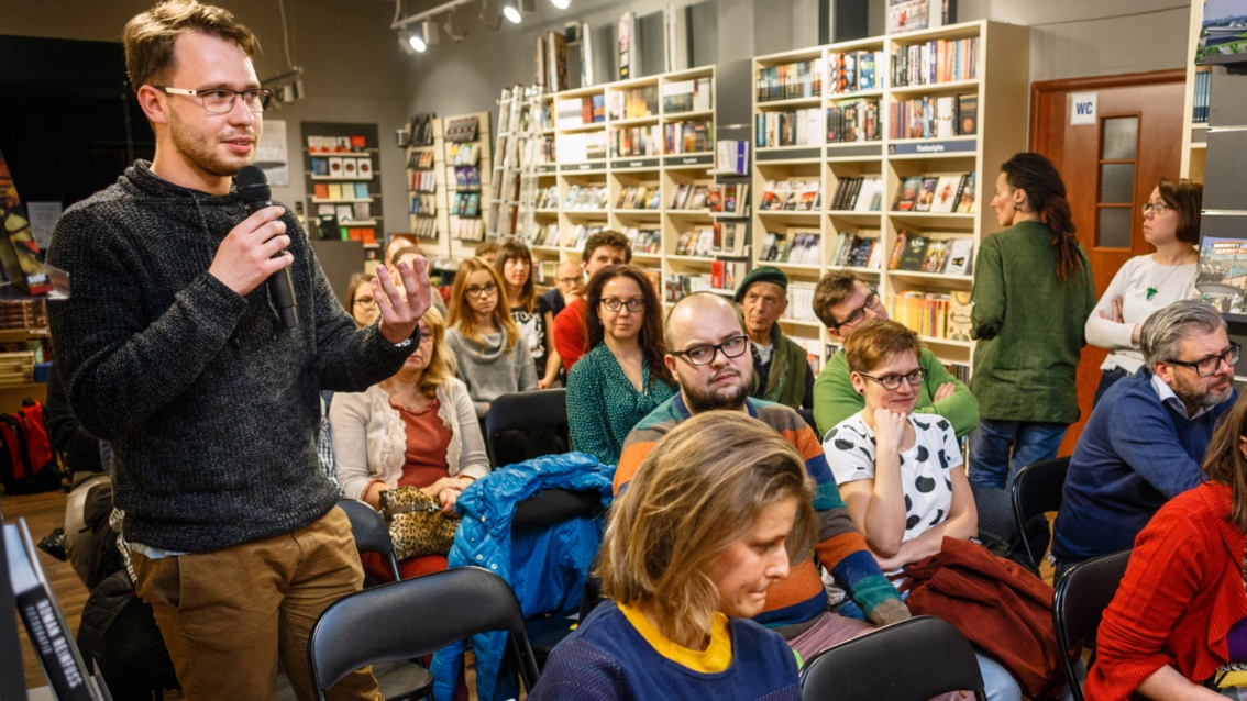 Publikum hört zu, ein Mann steht, hält ein Mikrofon in der Hand unn spricht, im Hintergrund volle Bücherregale