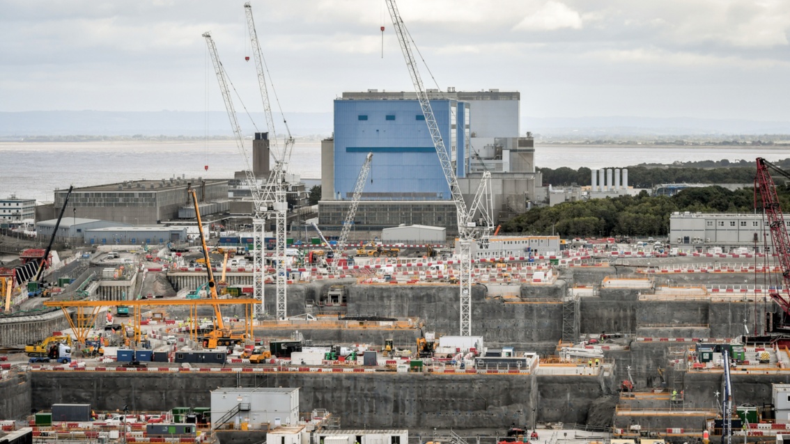 Große Baustelle mit Kränen und Betonmischer, im Hintergrund der blau bemalte Reaktorblock A des AKW HInkley Point