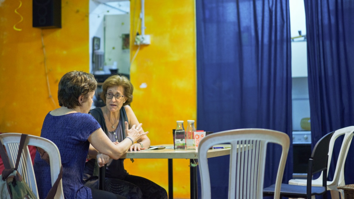 Ein Raum mit gelben Wänden und blauen Gardinen, zwei Frauen sitzen am Tisch und unterhalten sich.
