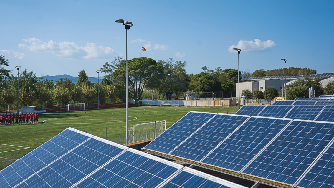 Fußballplatz, im Vordergrund Photovoltaikanlagen, im Hintergrund  das Spielfeld mit Flutlichtmasten