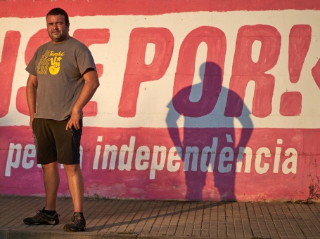 Ein Mann im T-Shirt steht vor einer Mauer, auf der der Slogan Sense Por! aufgemalt ist.
