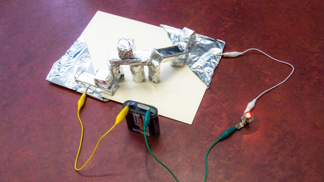 Das Experiment ist aufgebaut: Auf einer Pappe eine Konstruktion mit in Alufolien eingewickelten Bauklötzen; an den Seiten angeschlossene mit Kabel, wiederum verbunden mit einer Batterie und einer kleinen Glühbirne.