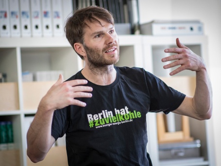 Ein junger Mann gestikuliert beim Sprechen; sein T-Shirt ziert das Logo der Kampagne «Berlin hat zu viel Kohle».