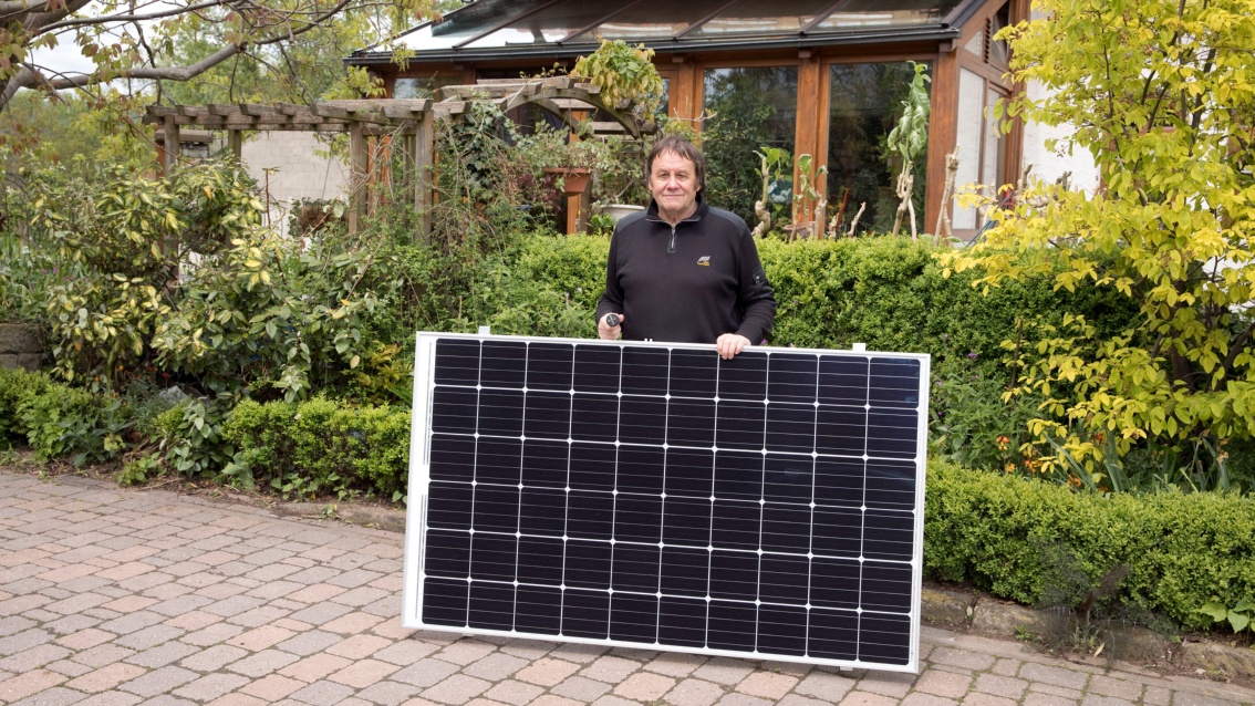 Ein Mann mittleren Alters präsentiert ein Solarmodul auf dem Gehweg vor seinem Einfamilienhaus.