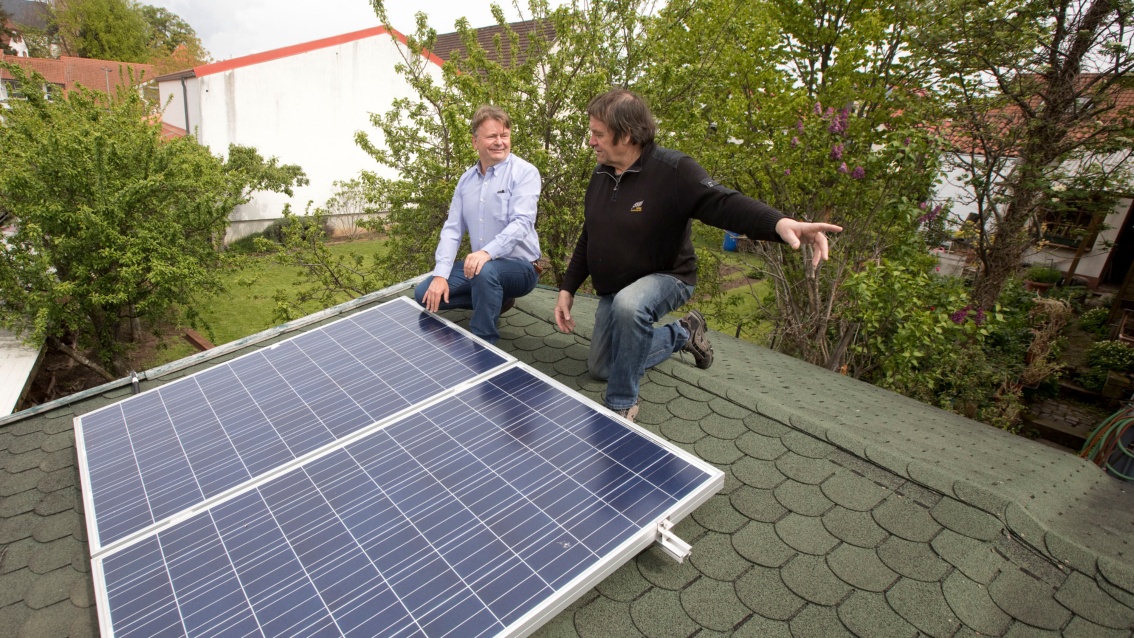 Zwei Männer knien auf dem Dach eines Gartenhäuschens neben einem PV-Modul, einer der beiden deutet nach  rechts.