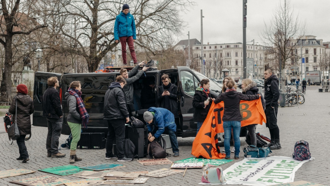 Zwölf junge Leute vor einem schwarzen Van, sortieren auf dem Boden liegende Demo-Plakate.