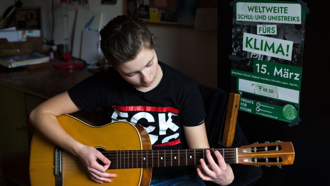 Ein junges Mädchen in einem T-Shirt mit dem Aufdruck: "FCK NZS" spielt auf einer akustischen Gitarre.