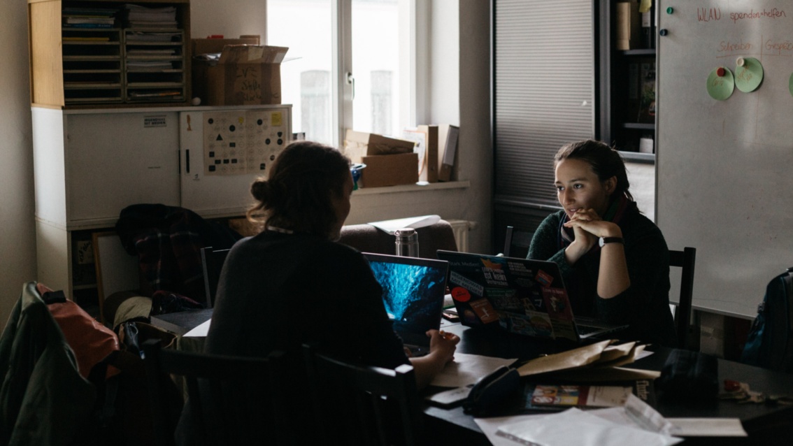 Zwei junge Frauen sitzen in einem dunkeln und etwas chaotischen Büro an einem Schreibtisch.