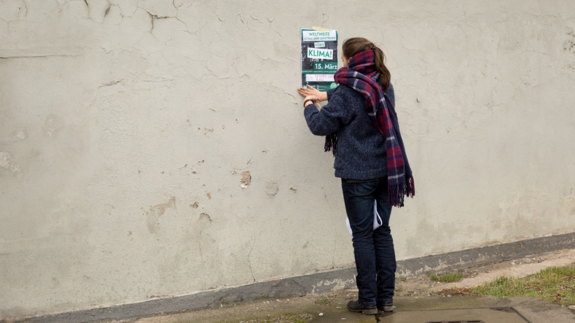 Eine junge Frau klebt ein Plakat an eine kahle Hauswand.