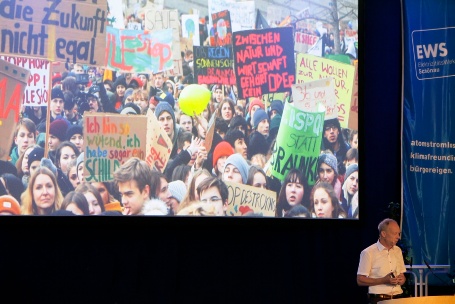 Ein dynamisch wirkender Mann mit Headset auf einer Bühne, hinter ihm eine Leinwand mit Bildern von einer Klimademonstration