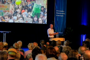 Man hält Vortrag, Publikum im Vordergrund und Leinwand mit Foto von Friday-for-Future-Demonstration im Hintergrund