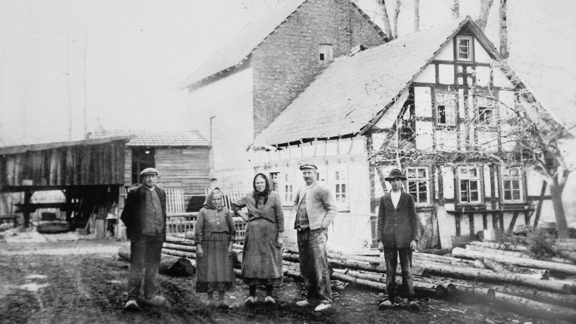 Historisches Foto von Personen vor einer Sägemühle, hinter ihnen liegen noch ungeschälte Baumstämme.