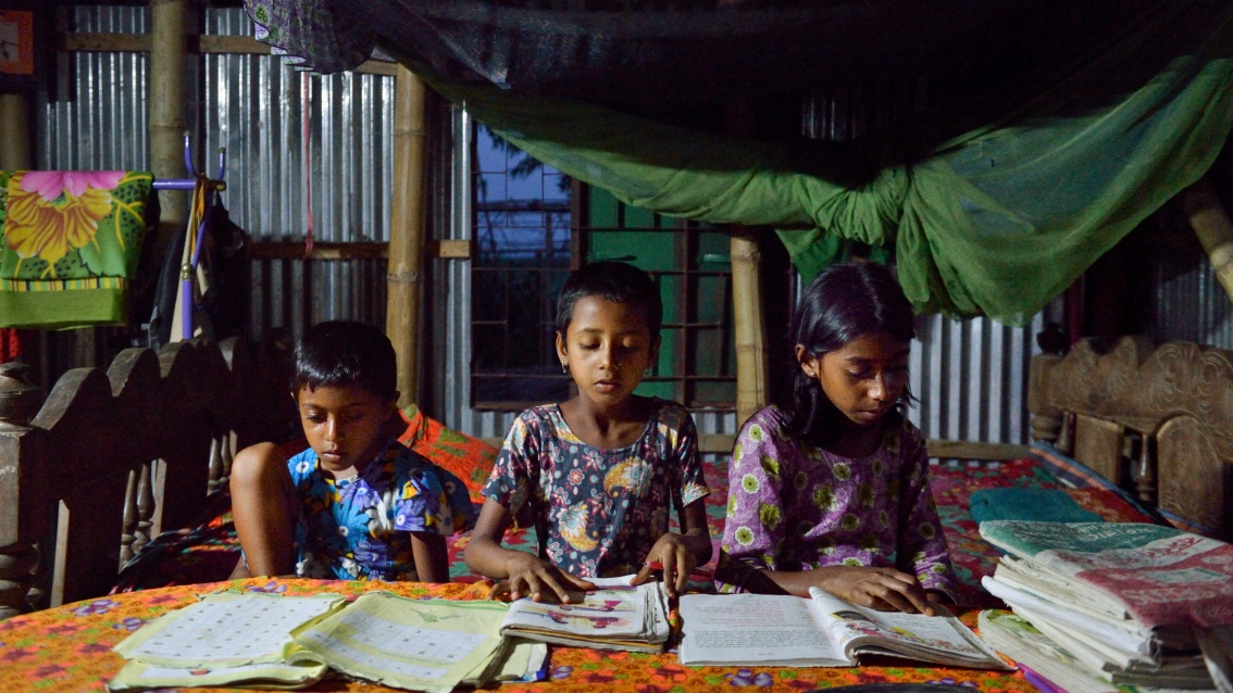 In einer einfachen Behausung sitzen drei Kinder vor Schulheften an einem Tisch, von oben scheint das fahle Licht einer Lampe.
