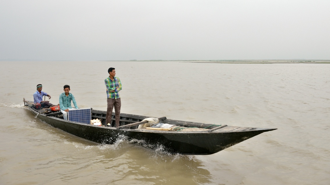 Auf einem sehr breiten Fluss, bei dunstigem Wetter, fährt ein schmales Boot mit drei Männern; geladen ist ein kleines Solarpanel.