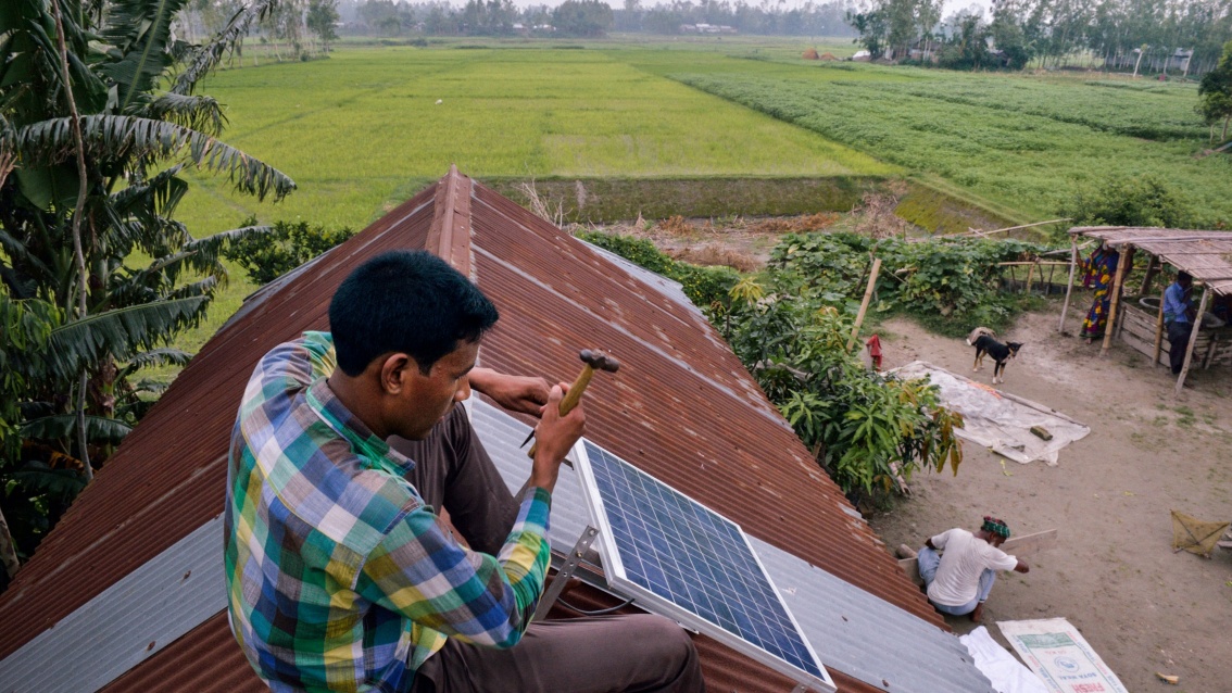 Auf einem Hausdach sitzt ein Mann und installiert ein kleines Solarpanel. Im Hintergrund sind grüne Felder zu sehen.