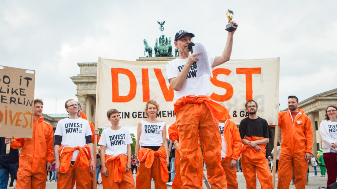 Vor dem Brandenburger Tor stehen Menschen in orangenen Overalls vor einem «Divest»-Transparent. Ein Mann spricht in ein Mikrofon und hält eine Art Pokal in die Luft.