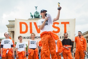 Vor dem Brandenburger Tor stehen Menschen in orangenen Overalls vor einem "Divest"-Transparent. Ein Mann spricht in eine Mikrofon und hält eine Art Pokal in die Luft.