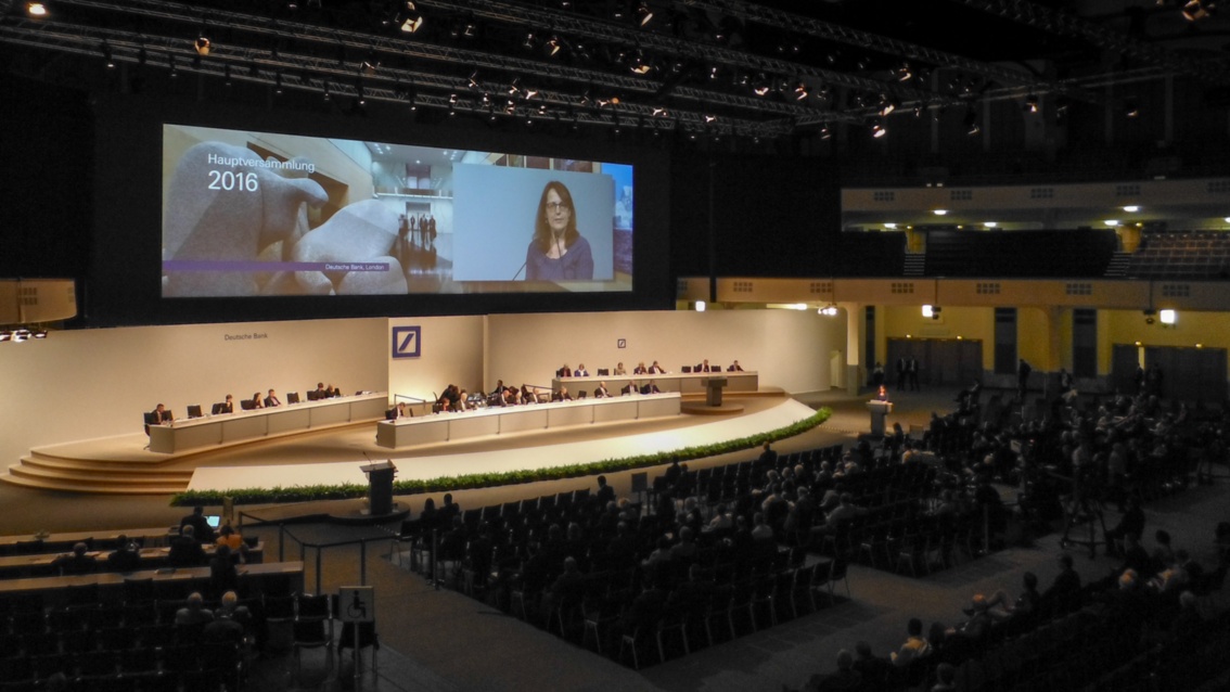 Ein riesiger Konferenzsaal mit sehr großer Bühne. Auf einer Großprojektion ist eine Frau am Mikrofon zu sehen.