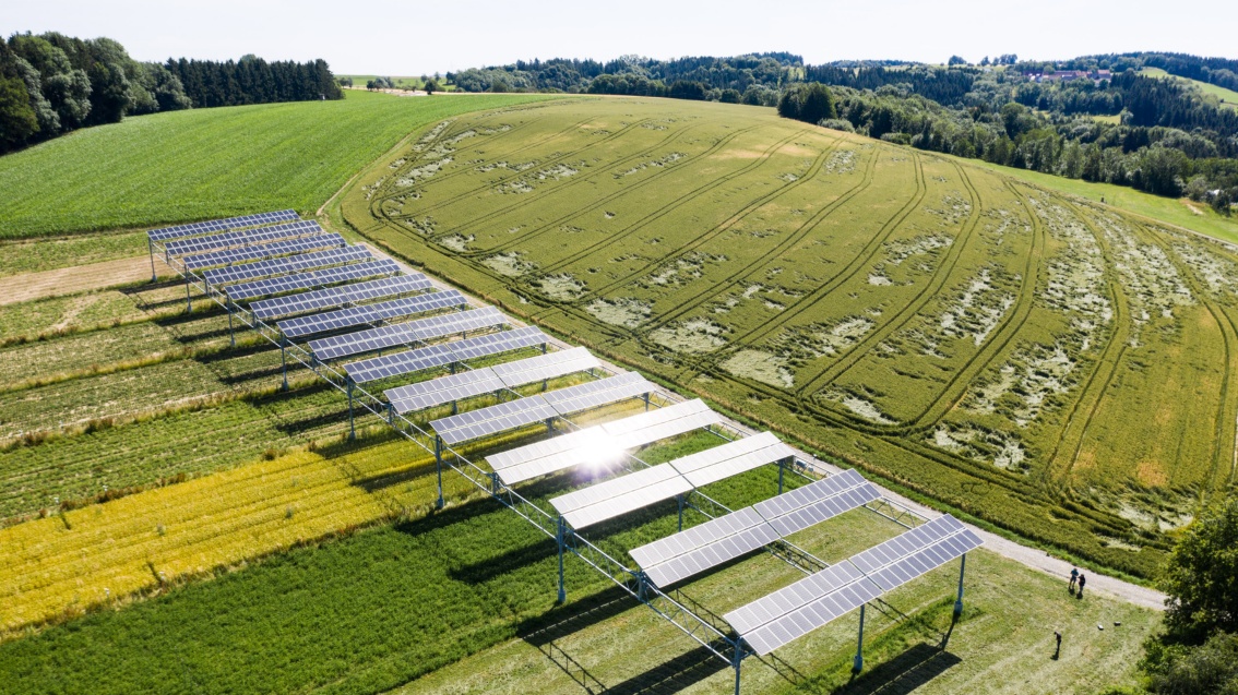 Aufnahme aus der Luft: Blick über eine hügelige Landschaft, im Vordergrund mehrere Reihen PV-Anlagen