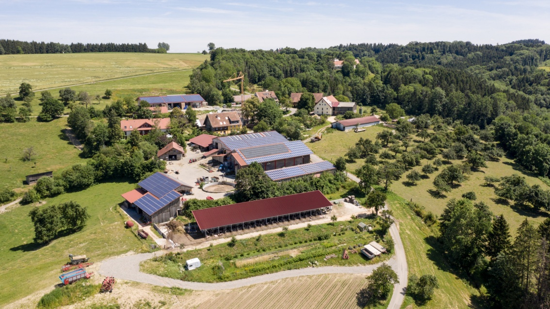 Luftaufnahme eines Bauernhofes in hügeliger Landschaft, auf einigen Dächern sind PV-Module installiert