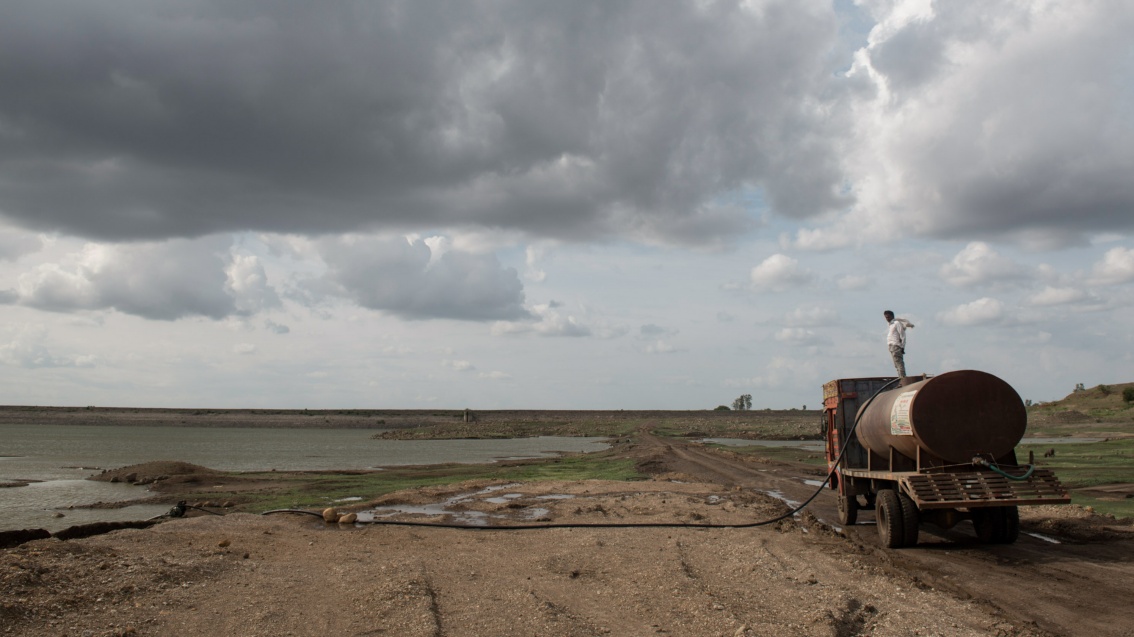 Ein Mann steht auf einem Tanklastwagen in einer öden flachen Landschaft, in der es nur noch ein paar, wie Pfützen anmutende Wasserstellen gibt.