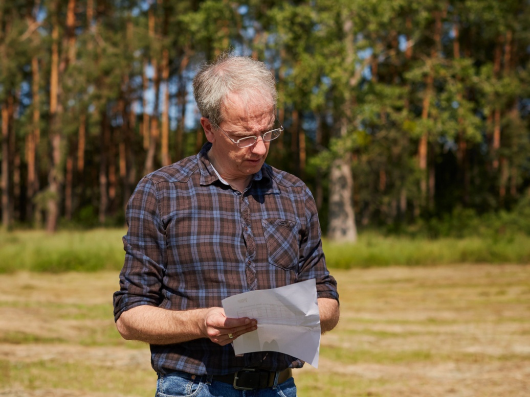  Ein grauhaariger Mann mit Brille, Karohemd und Jeans steht auf einer abgemähten Wiese und schaut auf einem Zettel; im Hintergrund ist ein lichter Kiefernwald zu erkennen.