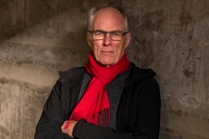 Älterer attraktiver Mann mit rotem Schal, draußen vor einer rostigen Wand fotografiert.
