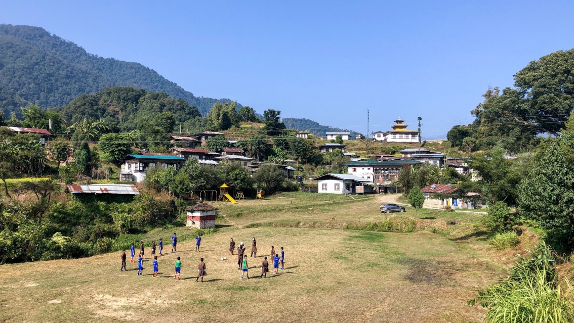 Auf einer großen Wiese stehen etwa 20 Kinder im Kreis, im Hintergrund sind Dorfgebäude vor bewaldeten Bergrücken zu erkennen.