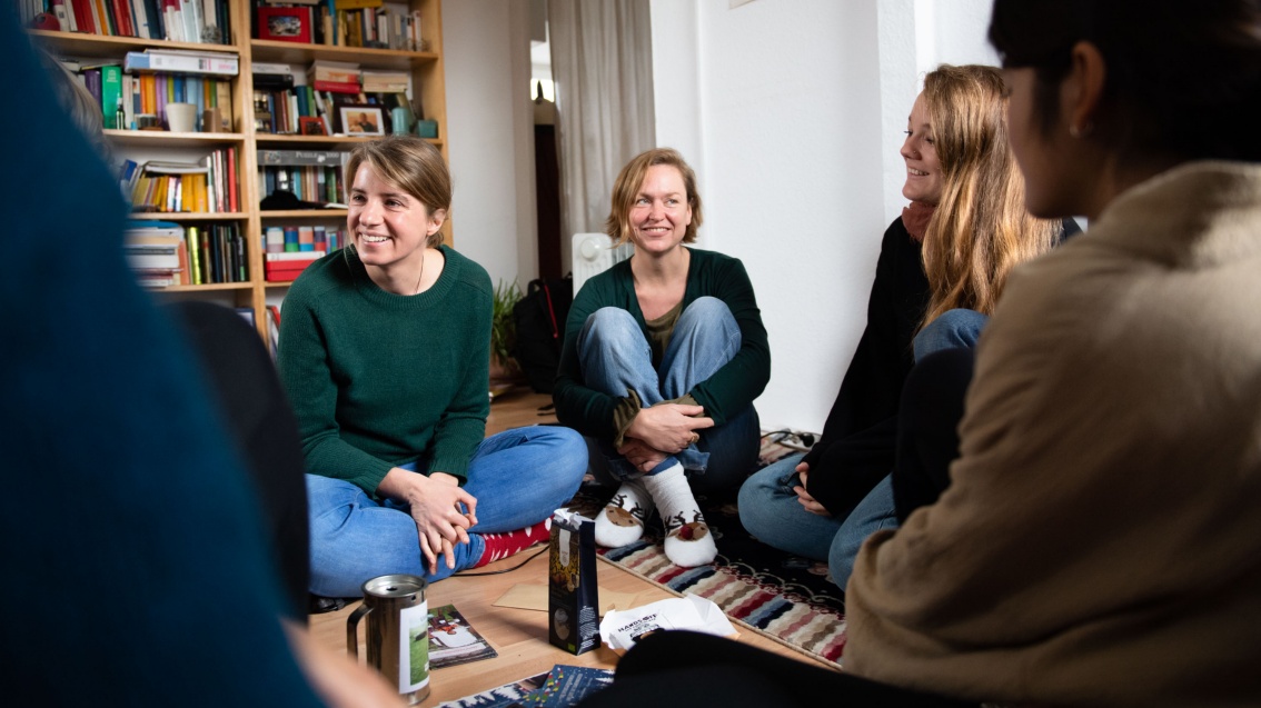 Fünf junge Frauen sitzen mit leger gekreuzten Beinen auf dem Boden eines Wohnzimmers.