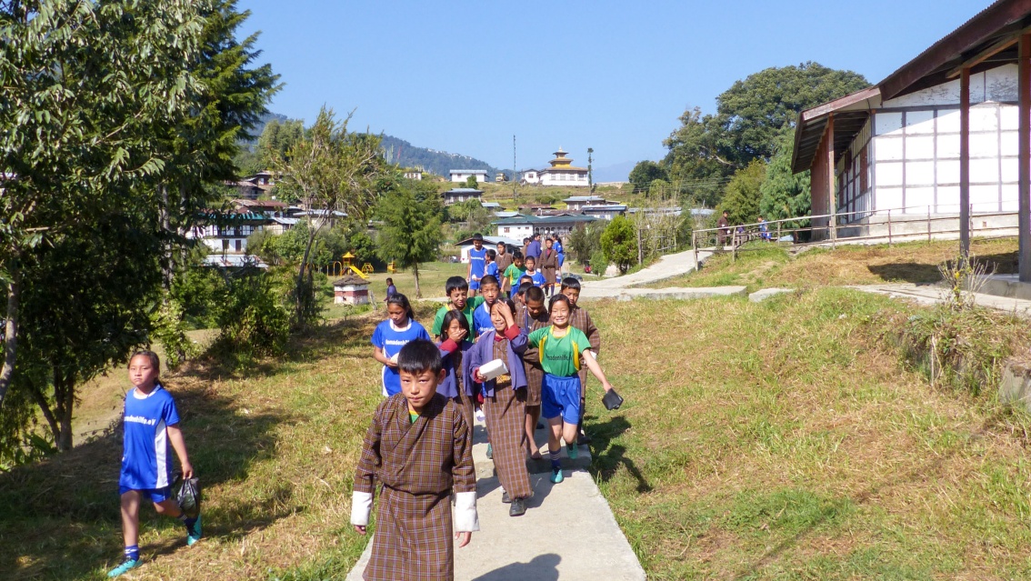 Eine Gruppe von Kindern, einige davon in Sporttrikots, laufen auf einem schmalen Weg auf den Fotografen zu, im HIntergrund Dorfgebäude.