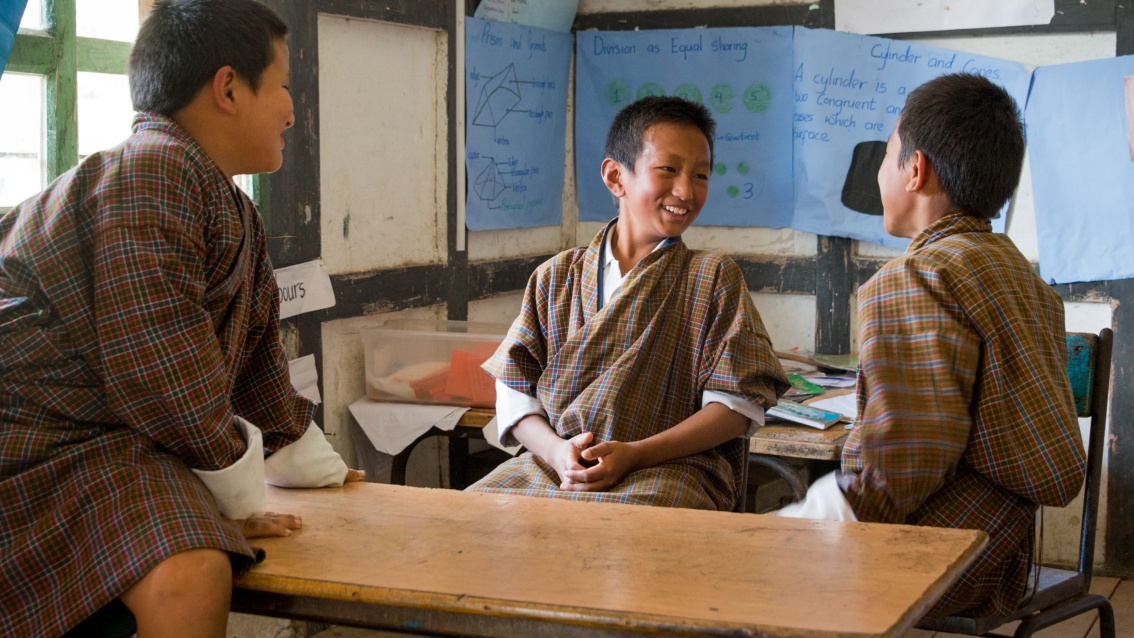 Drei Jungen um einen Schultisch plaudern lachend miteinander, im Hintergrund sind Papierbögen mit Schrift zu erkennen. 