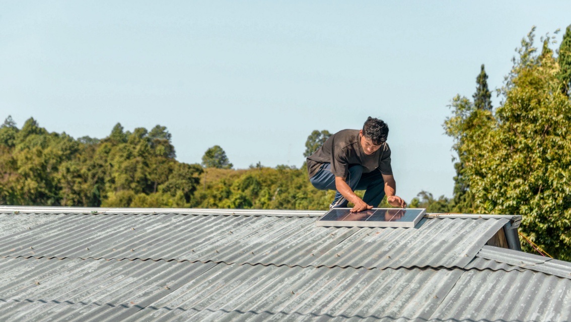 Ein junger Mann montiert in kniender Haltung ein kleines Solarmodul auf einem Wellblechdach.