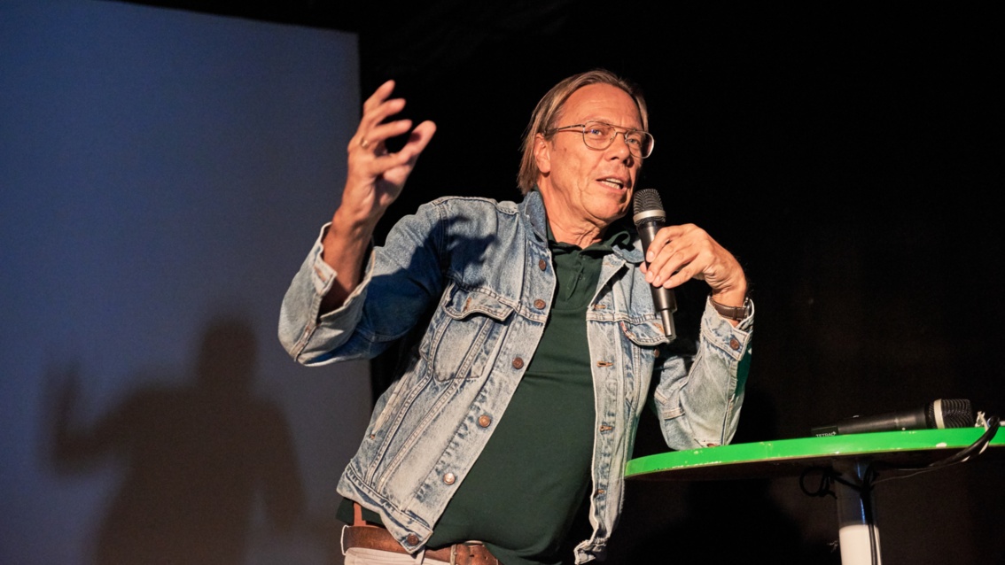 Ein älterer Mann in Jeansjacke auf der Bühne, locker auf einen Tisch gelehnt, spricht gestikulierend in ein Mikrofon.