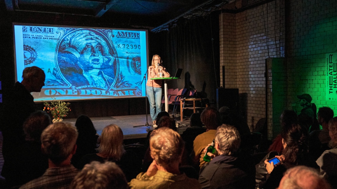 Blick aus dem Zuschauerraum auf die Bühne, wo eine Frau referiert. Auf der Projektionsfläche ist eine Dollarnote, auf der sich Roosevelt entsetzt die Haar rauft.