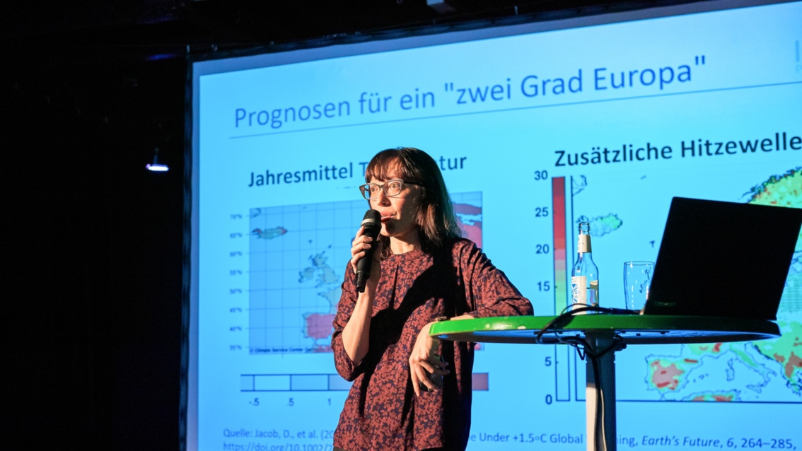 Eine junge Frau auf einer Bühne, aufgeklappter Laptop, im Hintergrund ein Schaubild, hält einen Vortrag.