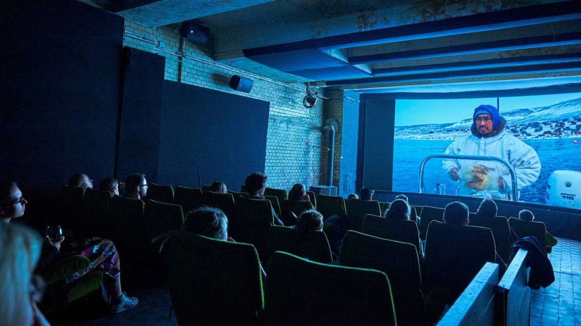 Eine halbvoll besetzter Kinosaal, vorne auf der Leinwand ist ein Inuit zu sehen, der in die Kamera spricht.