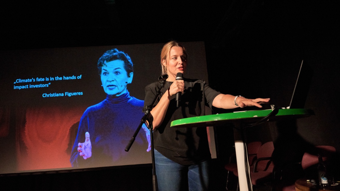 Eine junge Frau mit Mikrofon, referiert auf einer größeren Bühne. Im Hintergrund eine Projektion.