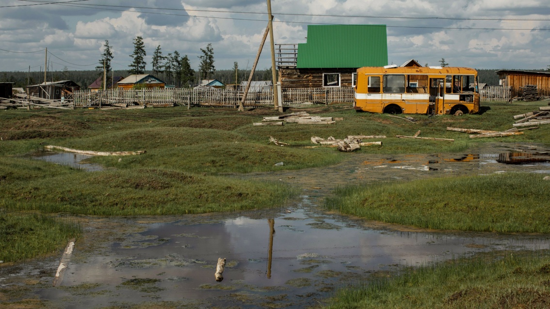Ein paar Häuser hinter einem Holzzaun, davor ein schrottreifer Bus, davor hügeliges Gras mit einer Wasserfläche