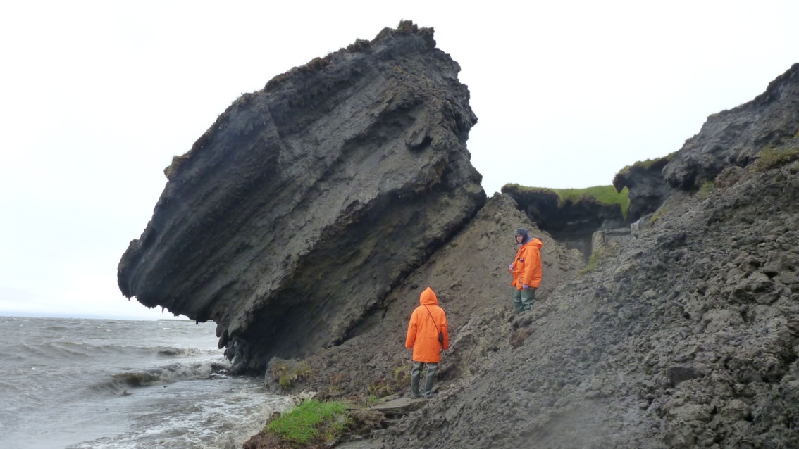 Zwei Männer in orangefarbenen Jacken an einer Küste. Vor ihnen  ein sehr großer Erdbrocken, der von der Uferkante abgebrochen ist.