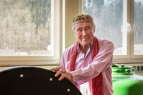 Ein freundlicher älterer Mann mit einer bestickten Kappe auf dem Kopf steht in einer Werkstatt.