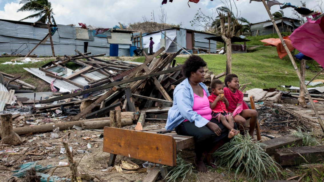 Eien Frau sitzt mit ihren 2 Kindern auf den Resten eine Bettes, draußen vor einem zerstörten Haus.
