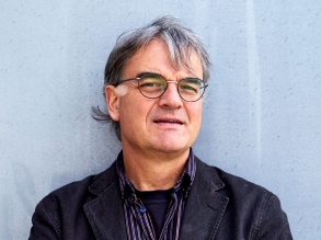 Mann mit grauen Haaren und Brille vor einer blauen Wand