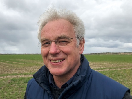 Portrait eines lächelnden Mannes mit grauen, windzerzausten Haaren vor einem Feld.