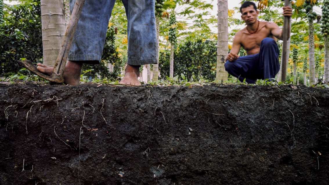 Ein Bodenquerschnitt zeigt unten den Humusboden, oben stehen zwei südamerikanische indigene Männer.
