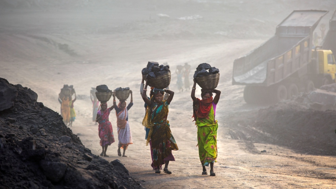 Zwischen Bergen von Kohle laufen indische Frauen mit Körben voller Kohlenstücken entlang.