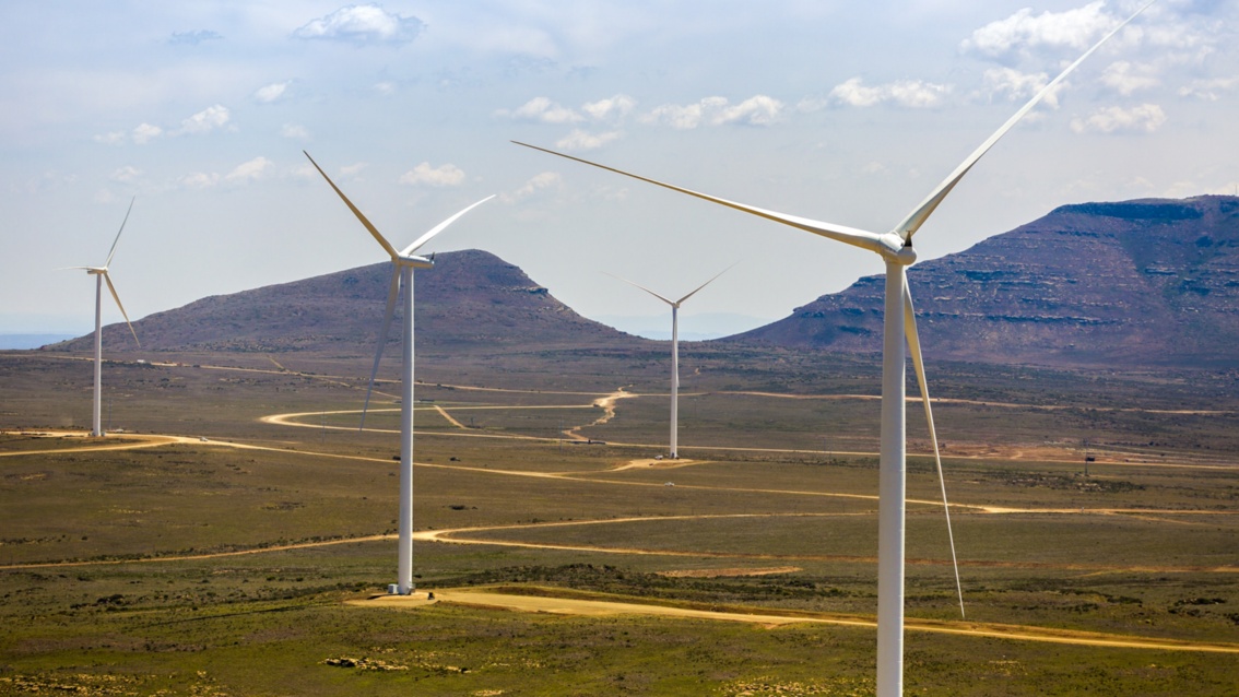 Vier große Windkraftanlagen in einer wüstenartigen Landschaft, im Hintergrund sind Berge zu erkennen.