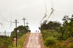 Ein aufsteigender Weg, gesäumt von Windrädern; zwei Menschen sind zentral am Horizont zu erkennen.